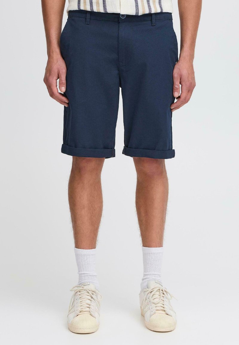 Mørkeblå bomullsshorts med smal passform, brettede manchetter og beltesløyfer. Kombinert med hvite treningssokker og beige joggesko.