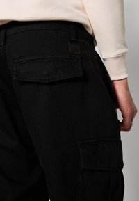 Pantalon cargo noir avec une finition texturée, doté de poches arrière et d'une petite étiquette. La ceinture est lisse, complétant le design général.