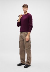 Bordeauxroter Strickpullover, khakifarbene Cargo-Hosen mit seitlichen Taschen und schwarze elegante Schuhe. Das Modell posiert mit einer Hand auf der Hüfte und der anderen Hand hält es eine Jacke.