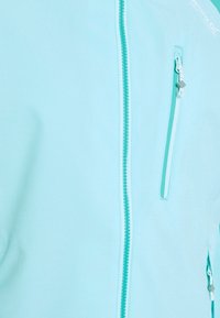 Veste bleu clair avec fermeture éclair à l'avant et poche poitrine verticale zippée, montrant la texture du tissu et les détails du curseur de la fermeture éclair.