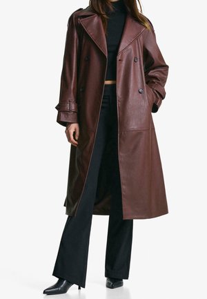 Mujer con un abrigo largo de cuero marrón, top negro, pantalones negros de pierna ancha y botas negras de tobillo con punta, de pie con una mano en el bolsillo.