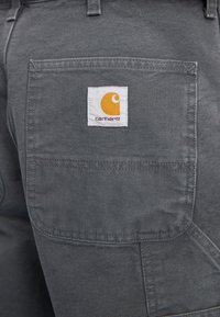 Primo piano di una tasca in tessuto grigio con una toppa del logo Carhartt in bianco, su cui è cucito un simbolo arancione.