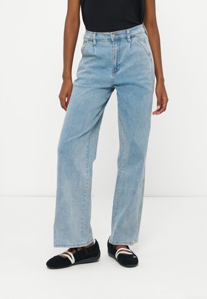 Femeie purtând blugi cu talie înaltă, drepti, albastru deschis, cu pliseuri, top negru și balerini negri cu bretele albe.