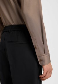 Lichtbruine, gestructureerde blouse met een ontspannen pasvorm en knoopmanchetten, gecombineerd met zwarte pantalons met elastische taille en een achterzak.