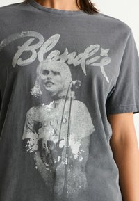 Harmaa graafinen T-paita, jossa on kulunut printti naisen muotokuvasta ja teksti "Blondie" tyylitellyllä fontilla. Lyhyet hihat, pyöreä kaula-aukko.
