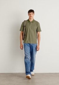 Olivengrünes Poloshirt mit Kragen, kombiniert mit hellblauen, geraden Jeans und weißen Sneakers. Braune Schultertasche getragen.