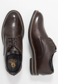 Une paire de chaussures habillées en cuir marron foncé, à lacets, avec des détails cousus et des semelles en caoutchouc sur fond blanc.