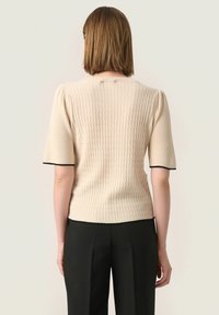 Beige svetr s texturovaným pleteným vzorem, krátkými rukávy a černým lemem na okrajích rukávů. Přiléhavý design s kulatým výstřihem.