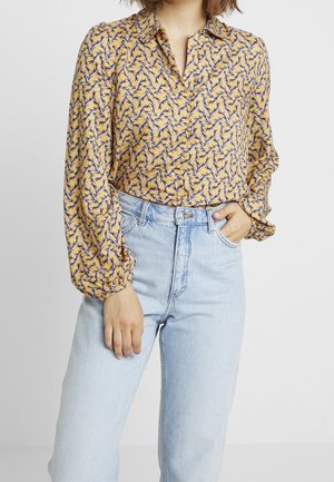 Žena v žluté košili s dlouhým rukávem s vzorem, zastrčenou do světle modrých džínů s vysokým pasem, ruka odpočívá v přední kapse.