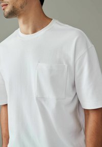 Hvid bomulds-T-shirt med rund halsudskæring, korte ærmer og en lomme på venstre bryst. Glat tekstur, afslappet pasform med minimale syninger.