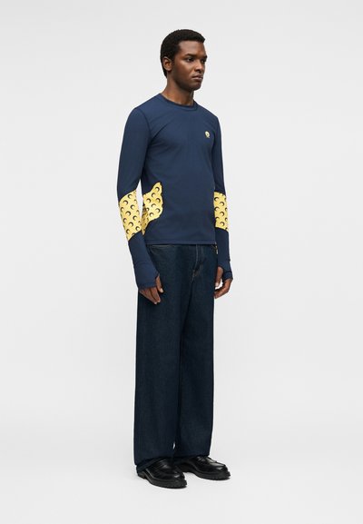 Άνδρας που φορά μπλε πουκάμισο με μακριά μανίκια και κίτρινα σχέδια σε σχήμα ημισελήνου στα μανίκια και σκούρο, φαρδύ τζιν, στέκεται μπροστά σε σκέτο φόντο.