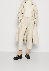 Beige trench coat with dark buttons, beige turtleneck, tailored cream trousers, and black loafers with gold chain accents. Patterned coat lining.