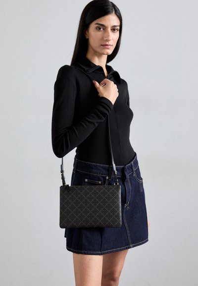Black crossbody bag with a geometric pattern, rectangular shape, and adjustable strap, worn with a long-sleeve top and denim skirt.