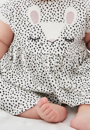 Niemowlę ubrane w biały strój z czarnymi kropkami i wzorem śpiącego zwierzątka, siedzące z widocznymi stopami na białej powierzchni.