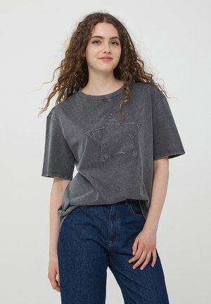T-shirt grigia oversize con un design a stella cucito sul davanti, maniche corte e scollatura rotonda, abbinata a jeans di denim blu.