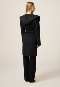 Cappotto nero su misura con cappuccio, cintura in vita arricciata e pantaloni neri a gamba larga, caratterizzati da una texture liscia e linee pulite.
