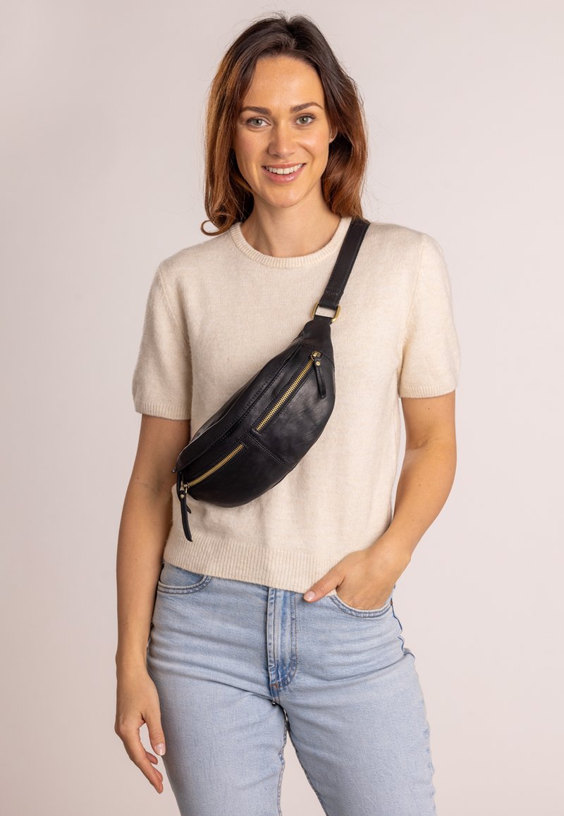 Schwarze Leder-Hüfttasche mit zwei Reißverschlusstaschen, geschwungener Form und vernickeltem Goldbeschlag. Quer über den Körper getragen, kombiniert mit einem leichten Pullover.