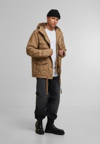 Parka marrón con capucha y forro de piel, que cuenta con múltiples bolsillos delanteros, cordones ajustables y un corte relajado, combinada con pantalones negros ajustados.