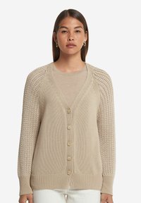 Beige stickad cardigan med djup V-ringning, femknapps stängning och vävda ärmar i texturerad spets. Mjuk textur och ribbade muddar.