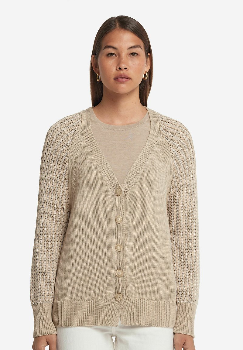 Beige stickad cardigan med djup V-ringning, femknapps stängning och vävda ärmar i texturerad spets. Mjuk textur och ribbade muddar.