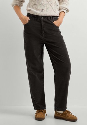 Person trägt schwarze Cordhose mit elastischem Bund, cremefarbenen Strickpullover und braune Freizeitschuhe mit Schnürsenkeln, steht mit den Händen in den Taschen.