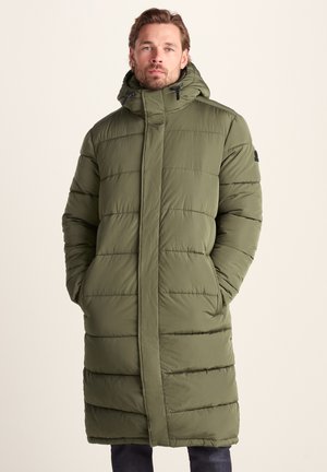 Man wearing long olive green padded winter coat with hood, hands in pockets, standing against plain light background.