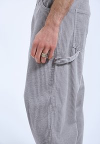 Grijze corduroy broek met verticale ribbels, zijzak, en een hand die een textuur ring draagt en de stof vasthoudt.