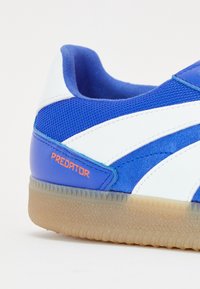 Zapatilla de deporte azul y blanca con el texto "PREDATOR" en naranja, parte superior de malla y ante, y suela de goma translúcida texturizada.