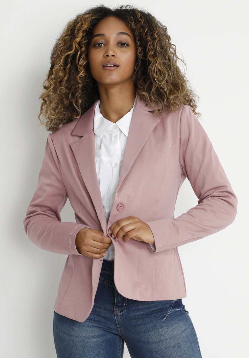 Blazer ajusté rose clair avec des manches longues et une fermeture à un bouton, associé à une chemise blanche à col. Texture de tissu lisse, design sur mesure.