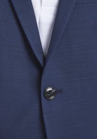 Veston de costume bleu marine boutonné sur une chemise blanche texturée, montrant le revers et un seul bouton noir.