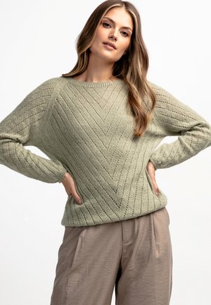 Jasnozielony sweter z dzianiny z wzorem w zygzak, okrągłym dekoltem i długimi rękawami, zestawiony z jasnobeżowymi spodniami.