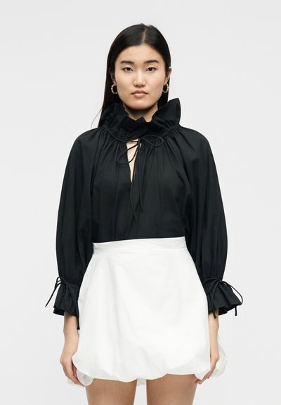 Ženska, ki nosi črno dolgoročno bluzo z gubanim ovratnikom in manšetami, kombinirano z belo mini krilico z naborki, stoji pred enobarvnim ozadjem.