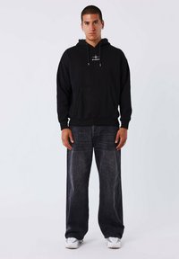 Hoodie nera con tasca frontale e logo bianco; abbinato a jeans scuri dalla vestibilità ampia e sneakers bianche. Design semplice, stile casual.