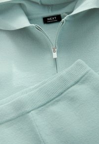 Ljusblå zip-up hoodie med ribbade ärmslut och nederkant, som har en metalldragkedja och en etikett med texten "NEXT, MADE IN MYANMAR."