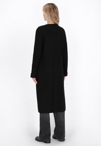 Donna con capelli biondi che indossa un lungo cardigan nero a coste, jeans a gamba larga scuri e scarpe nere, in piedi davanti a uno sfondo bianco, vista posteriore.
