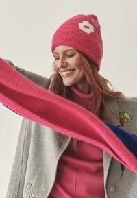 Pink knit beanie with white flower detail; model wearing a pink sweater and gray coat with embroidered accents, holding a multicolored scarf.