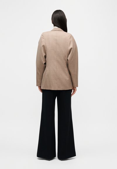 Blazer marron sur mesure avec épaulettes, fente unique et coupe décontractée, associé à un pantalon noir à jambes larges.