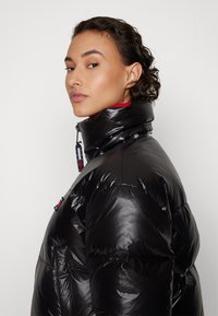Chaqueta de plumas negra con cuello alto, textura brillante y forro rojo. Incluye cierres de cremallera y botones a presión con acentos de marca en contraste.