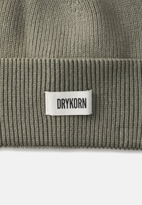 Horgolt sapka tompa olíva zöld színben, bordázott textúrával. Jellemzője egy fehér címke, amelyen a "DRYKORN" felirat található a márkázás érdekében.