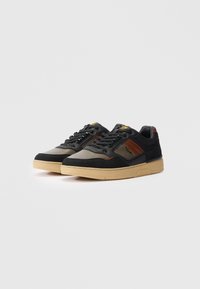 Un paio di sneaker basse con pannelli neri, marroni e grigi e suole beige, esposte su uno sfondo bianco puro.