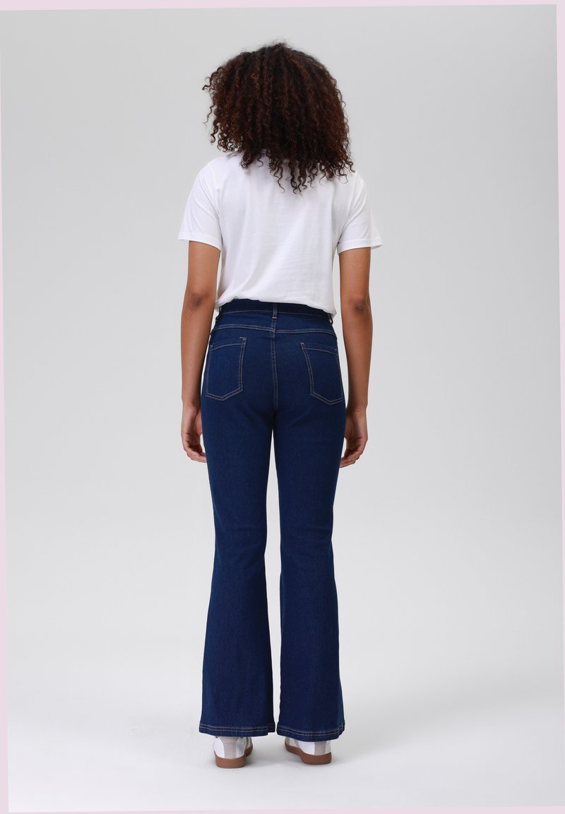 Jeans a zampa d'elefante blu con cuciture sbiadite, vita alta e due tasche posteriori. Indossati con una maglietta bianca corta e scarpe bianche.