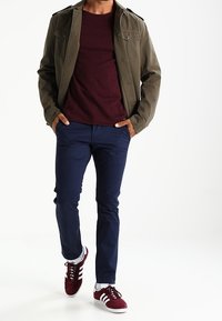 Olivgrüne Jacke, burgunderfarbenes Hemd, marineblaue Hose und rote Sneakers mit weißen Akzenten. Ausgestattet mit Taschen und einem klassischen Design.