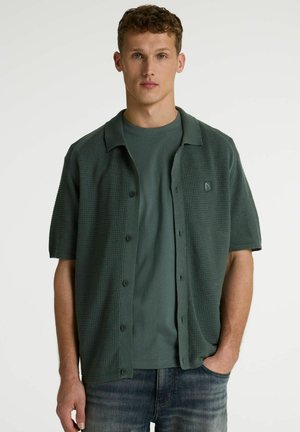 Junger Mann steht, trägt ein dunkelgrünes Strickhemd mit Waffelmuster und kurzen Ärmeln über einem passenden T-Shirt und blauen Jeans, Hand in der Tasche.