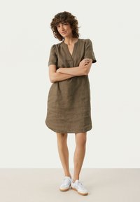 Olivgrünes Leinenkleid mit kurzen Ärmeln, V-Ausschnitt und lockerer Passform. Getragen mit weißen Sneakers, knielang mit abgerundetem Saum.