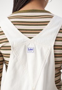 Personne portant une salopette de travail blanche Lee par-dessus une chemise à manches longues avec des rayures horizontales vertes, marron et beige, vue de dos.