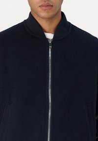 Tumedast villast bomber-jakk, millel on lukuga esikülg, soonikkoes krae ja küljetaskud. Sile tekstuur ja minimalistlik disain, ilma nähtavate logo'teta.