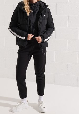 Svart vadderad jacka med huva, med quiltad textur och vita zickzack-accenter på ärmarna, kombinerad med svarta joggers och vita sneakers.