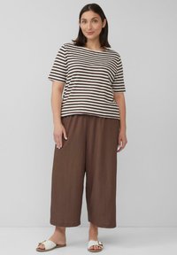 Gestreiftes braunes und cremefarbenes T-Shirt mit kurzen Ärmeln, kombiniert mit lockeren braunen weit geschnittenen Hosen. Das Model trägt cremefarbene Riemchensandalen mit einem einfachen Schnallen-Detail.