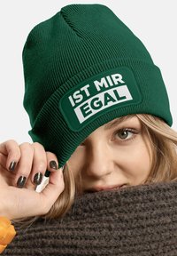 Green ribbed knit beanie with a rectangular white patch displaying the text "IST MIR EGAL." The wearer partially hides behind a dark knitted scarf.