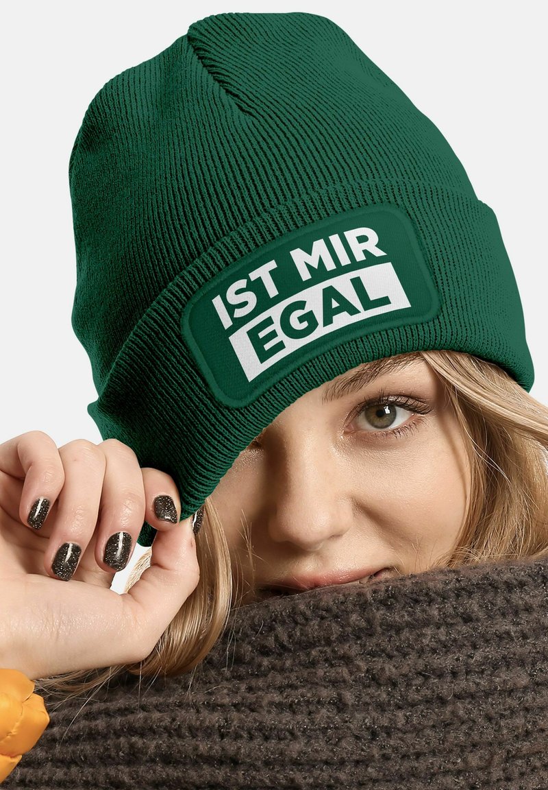 Green ribbed knit beanie with a rectangular white patch displaying the text "IST MIR EGAL." The wearer partially hides behind a dark knitted scarf.